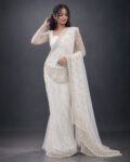 A woman wearing an elegant white and gold bridal saree with intricate embroidery, lace borders, and delicate floral detailing, posing in a studio