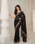 A woman wearing an elegant black saree with intricate gold floral embroidery and a delicate embellished border, posing in a luxurious indoor setting